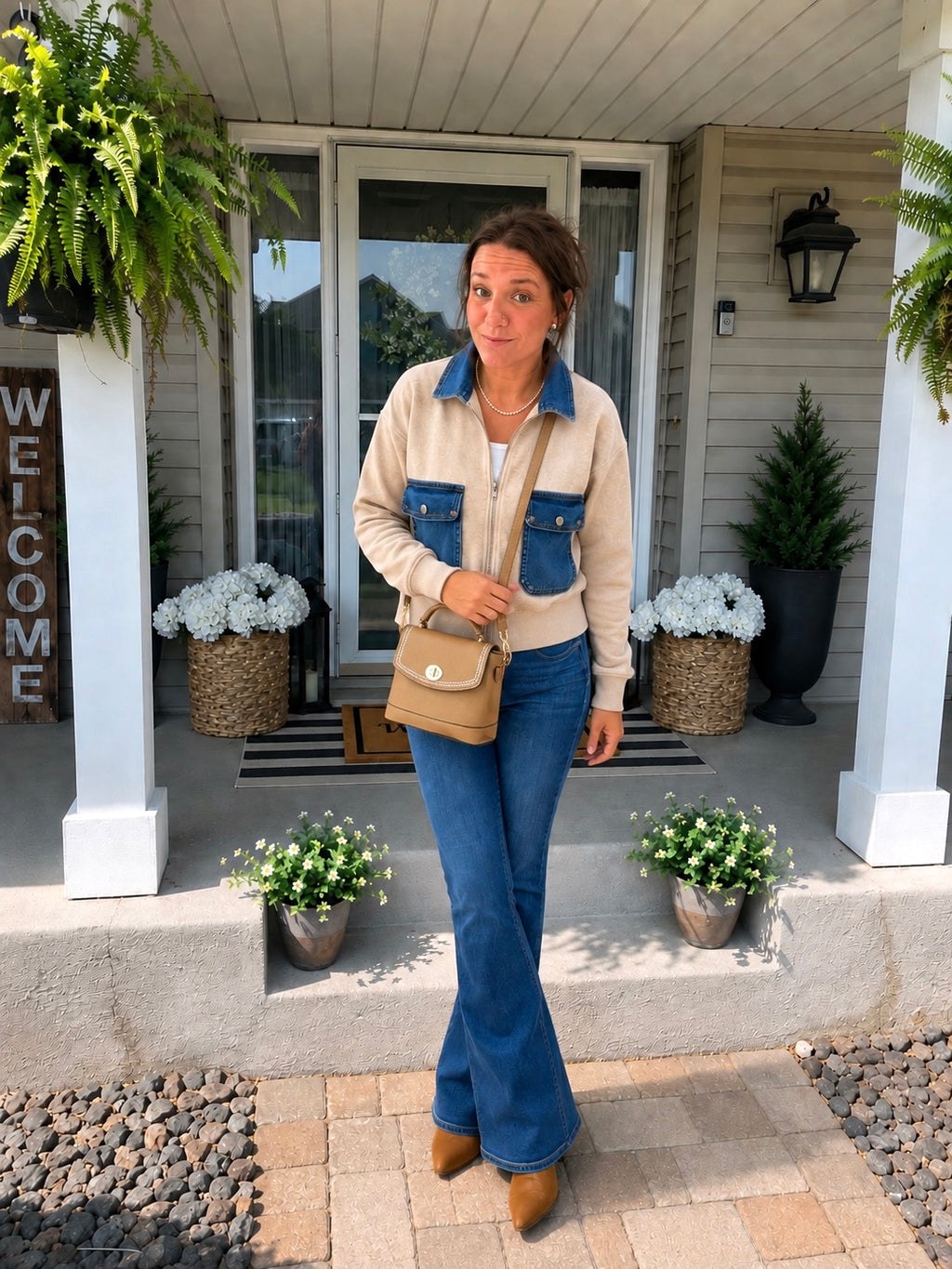 A woman stands on a front porch wearing a beige and blue cropped jacket, blue jeans, and brown boots, while carrying a tan shoulder bag. The porch has white columns, potted plants, and a welcome sign.