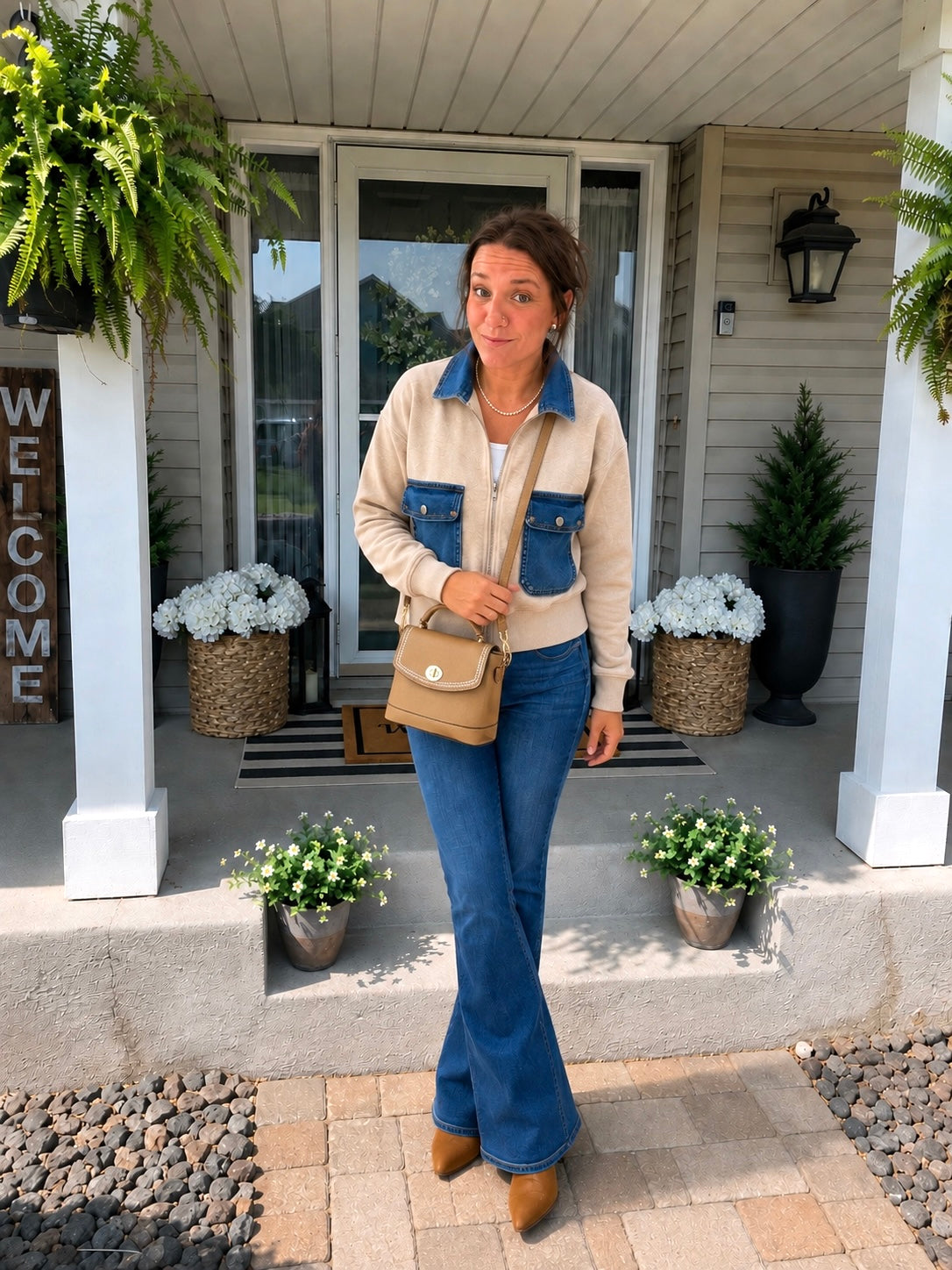 A woman stands on a front porch wearing a beige and blue cropped jacket, blue jeans, and brown boots, while carrying a tan shoulder bag. The porch has white columns, potted plants, and a welcome sign.