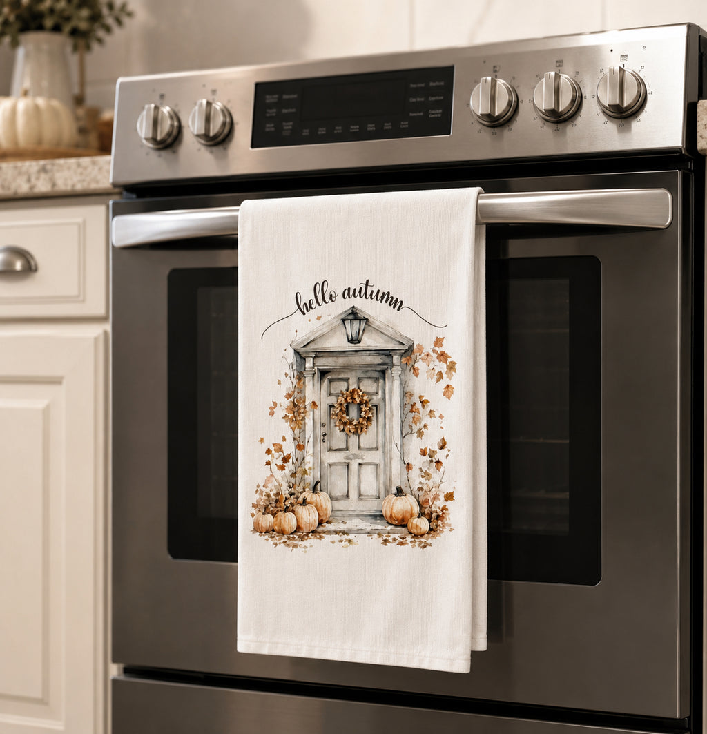 A white kitchen towel with an autumn-themed illustration of a front door, pumpkins, and the words 'hello autumn' hanging on the handle of a stainless steel oven in a kitchen.