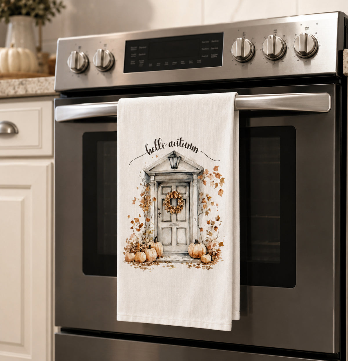 A white kitchen towel with an autumn-themed illustration of a front door, pumpkins, and the words 'hello autumn' hanging on the handle of a stainless steel oven in a kitchen.