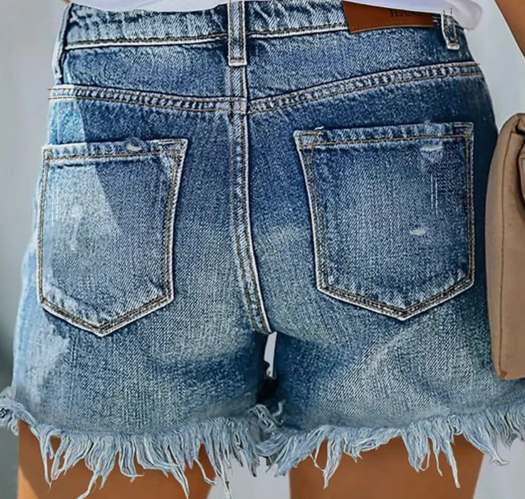 Close-up back view of light blue denim shorts with a raw frayed hem, featuring two large patch pockets and a brown leather waistband patch.