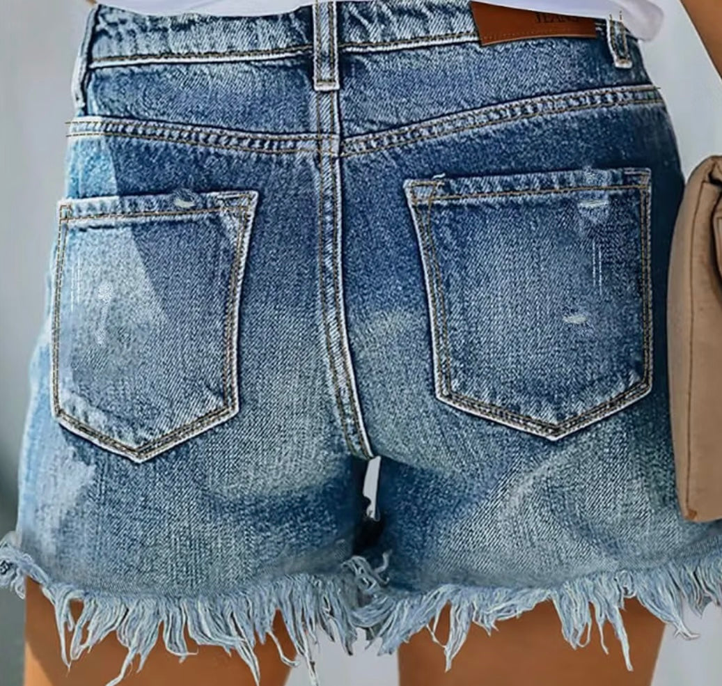 Close-up back view of light blue denim shorts with a raw frayed hem, featuring two large patch pockets and a brown leather waistband patch.