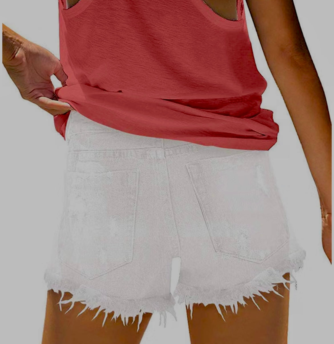 White distressed denim shorts with a raw frayed hem, shown on a model wearing a coral red tank top against a solid gray background.