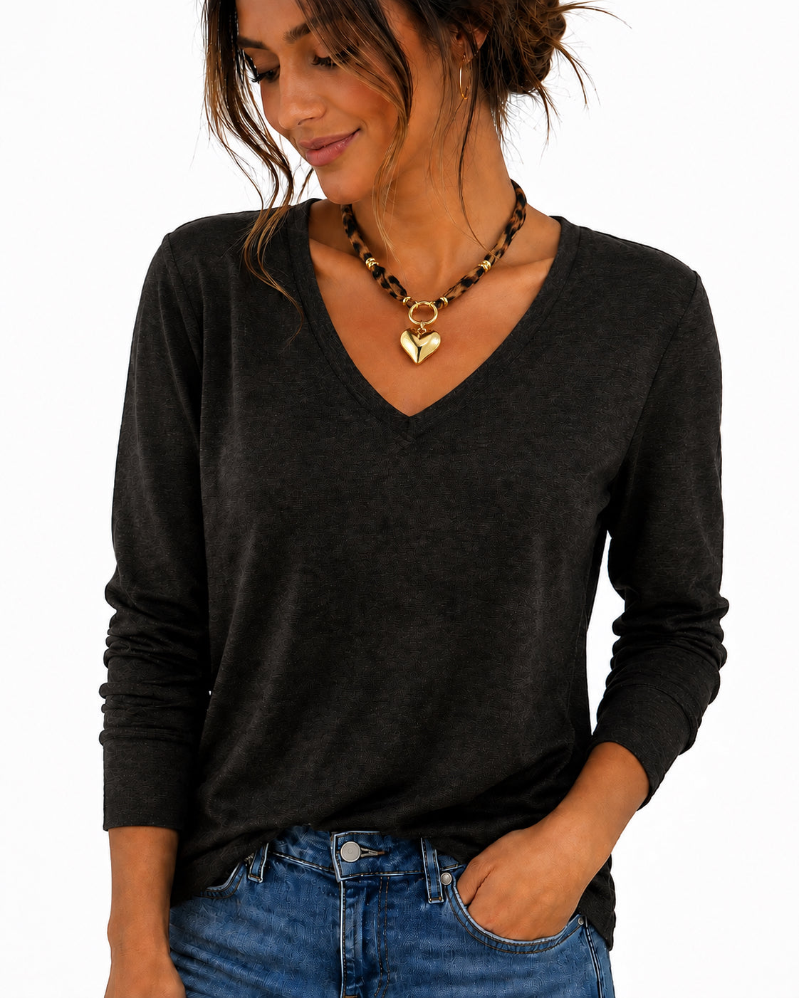 A woman wearing a dark charcoal gray long-sleeve V-neck t-shirt and blue jeans, accessorized with a gold heart pendant necklace, against a plain white background.