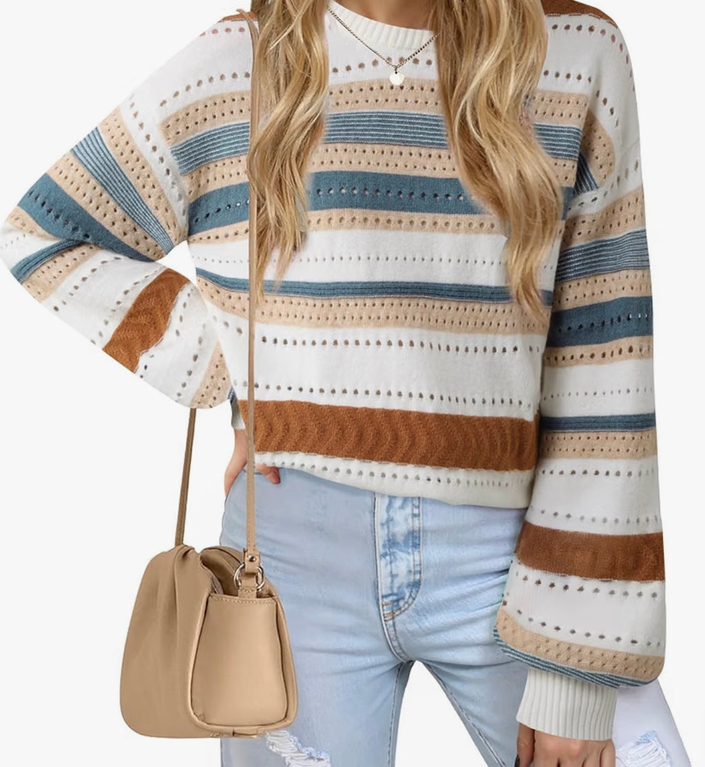A woman wearing a cream, blue, and brown striped knit sweater with long sleeves, paired with light blue jeans and a beige crossbody bag, shown against a plain white background.