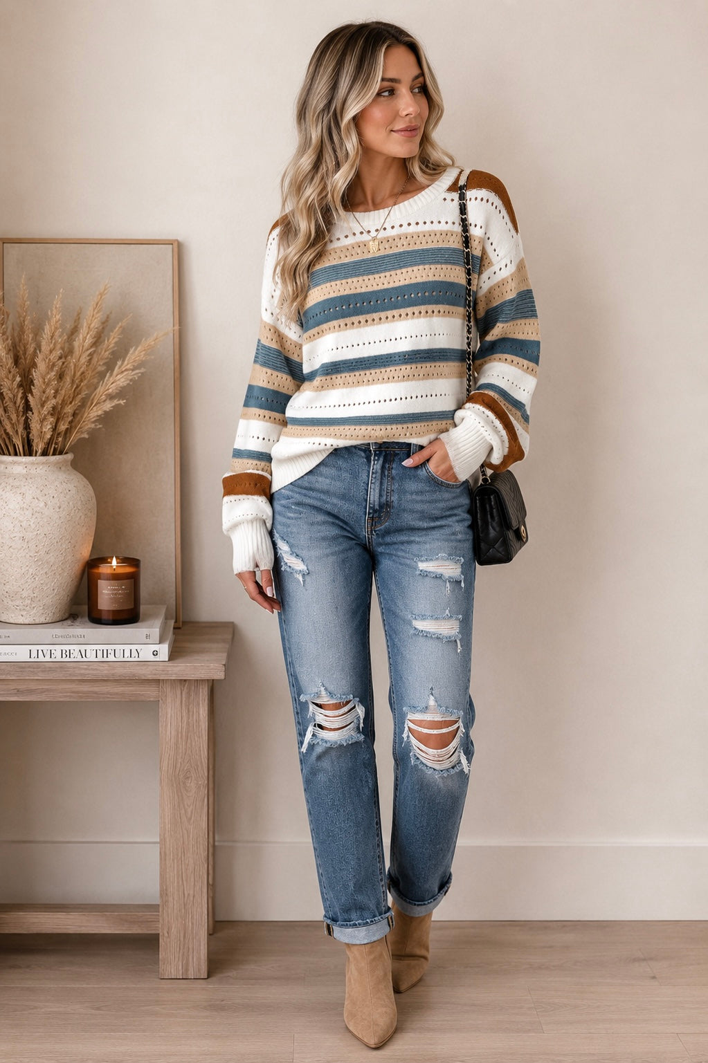 A woman stands indoors wearing a knit sweater with horizontal stripes in cream, tan, and muted blue, paired with distressed blue jeans and tan ankle boots. She is posed in a styled room with a side table, dried grass in a vase, and a candle.