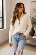 A woman wearing a cream cable-knit quarter-zip sweater and light-wash distressed jeans, standing in a bright, modern room.