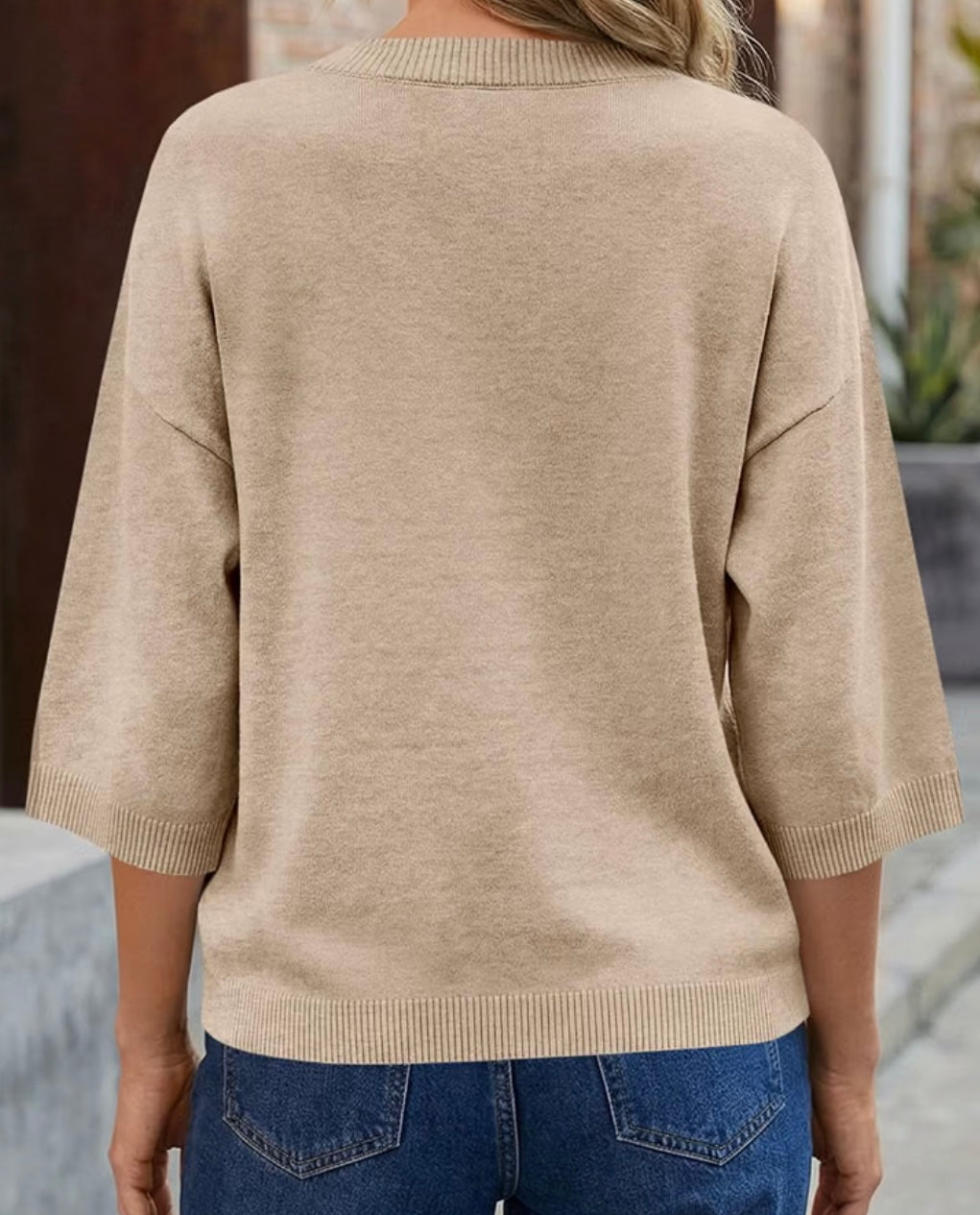 A woman viewed from the back wearing a light beige knit sweater with three-quarter sleeves and blue jeans.
