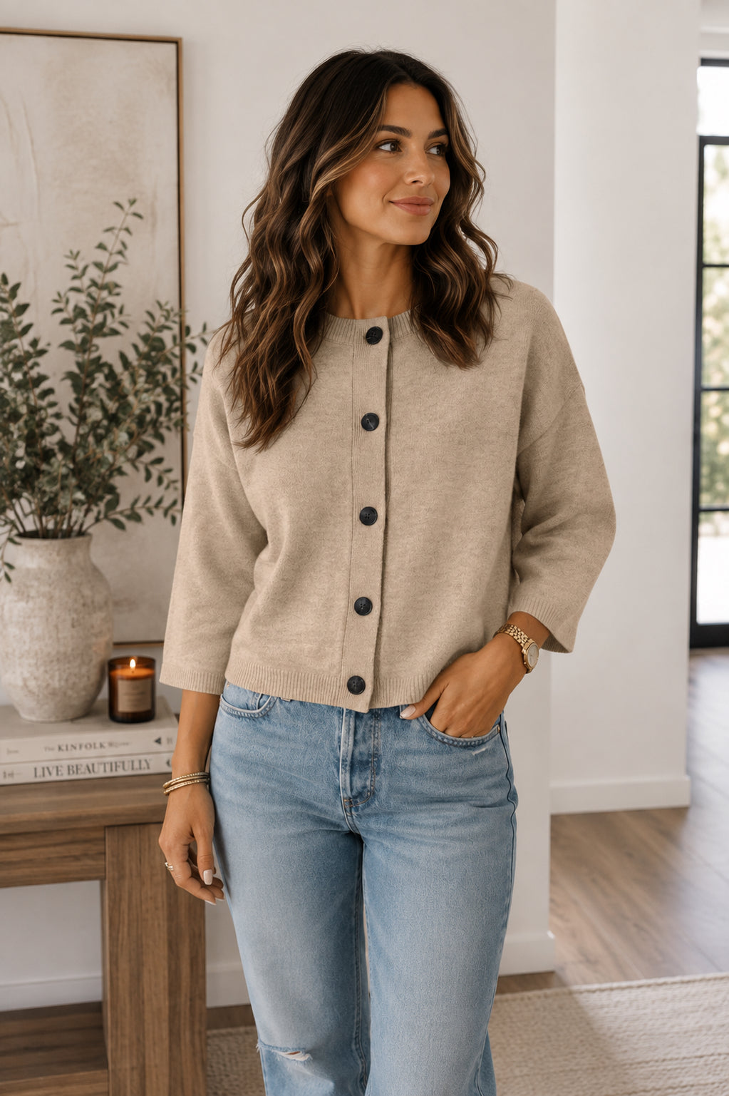 A woman wearing a beige cropped button-front cardigan with three-quarter sleeves and light-wash blue jeans, standing in a bright, modern room with a wooden console table and framed wall art.