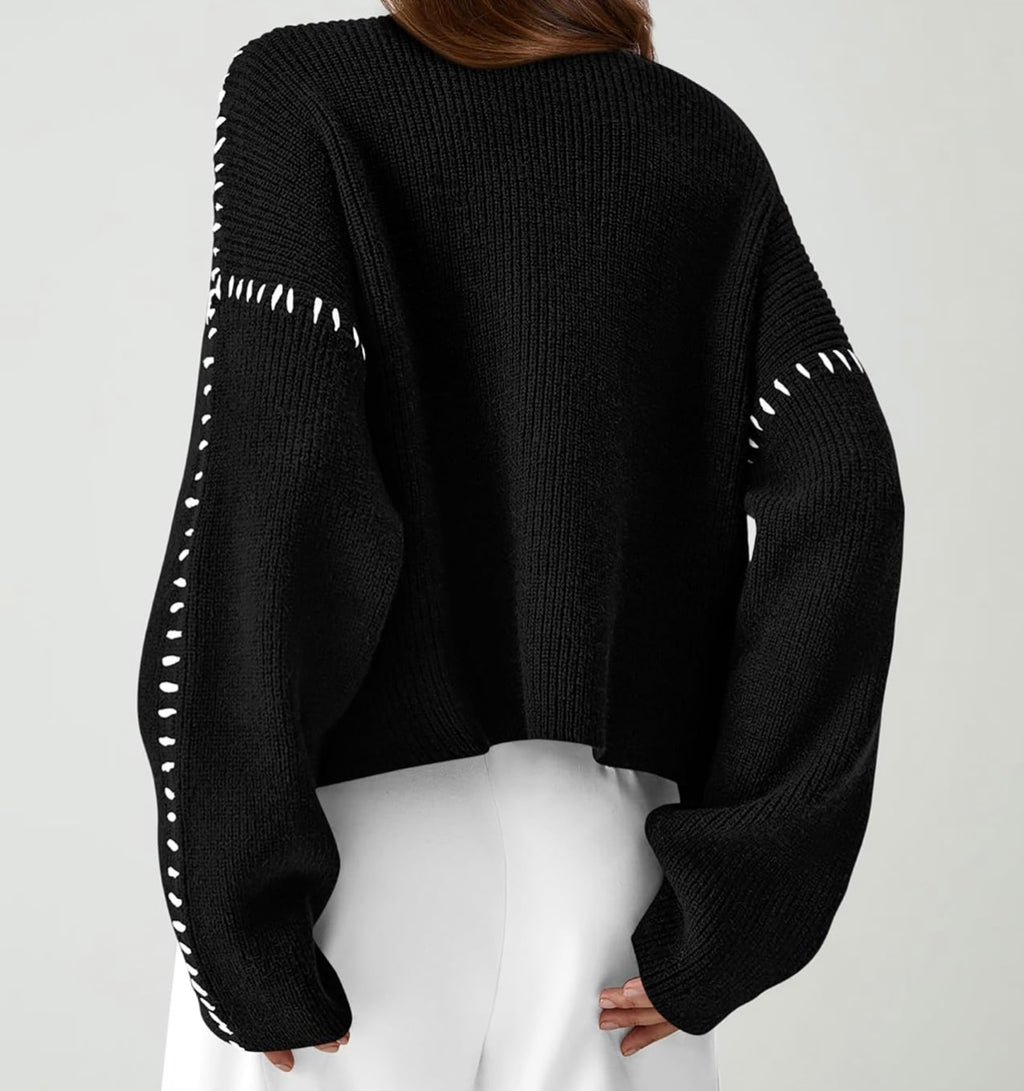 A person shown from the back wearing a black ribbed knit cropped sweater with white contrast stitching details along the sleeves, paired with white pants, against a light gray studio background.