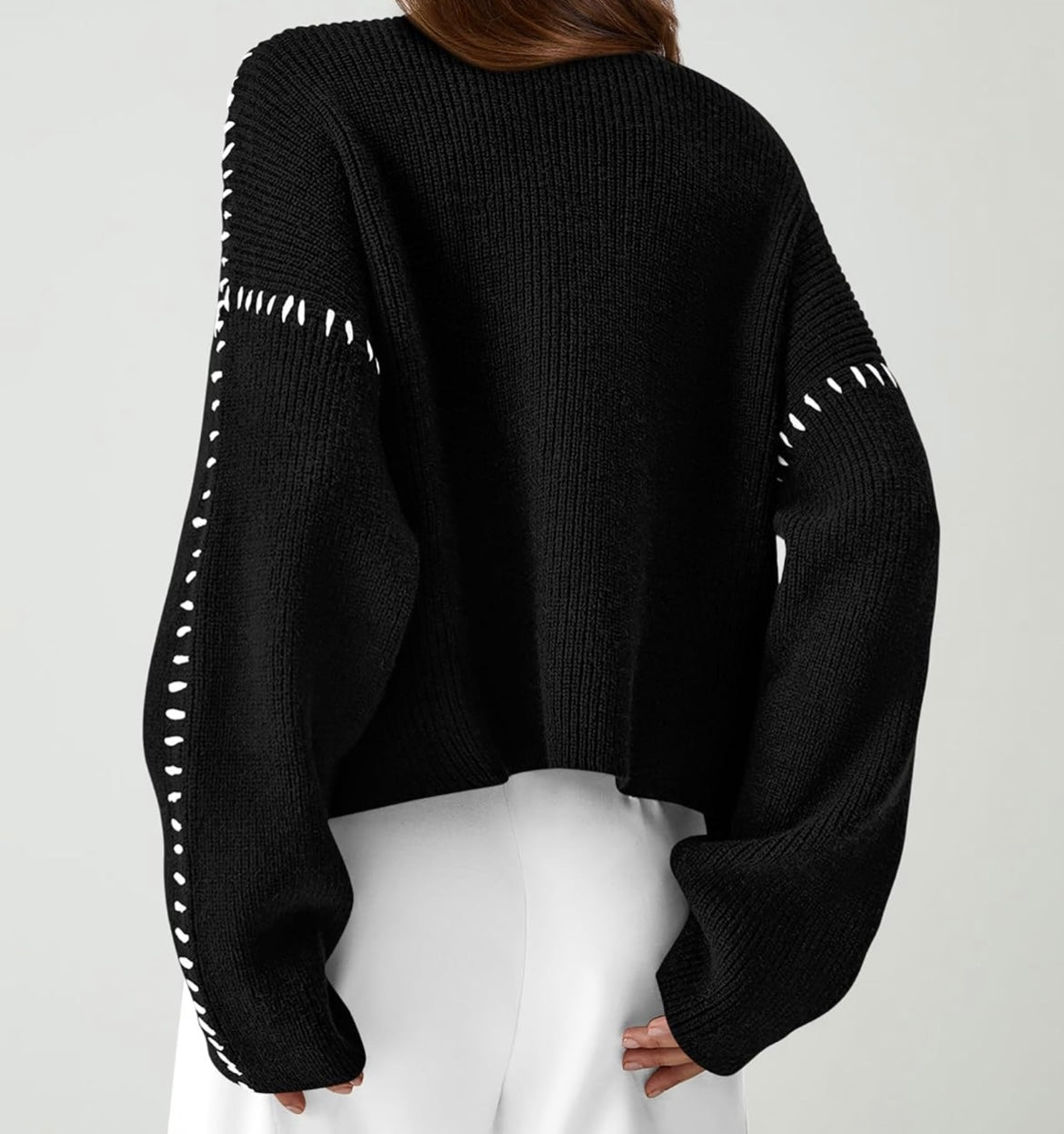 A person shown from the back wearing a black ribbed knit cropped sweater with white contrast stitching details along the sleeves, paired with white pants, against a light gray studio background.