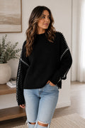 A woman wearing a black ribbed knit sweater with white dotted trim along the sleeves, paired with light blue ripped jeans, standing in a bright, modern room.