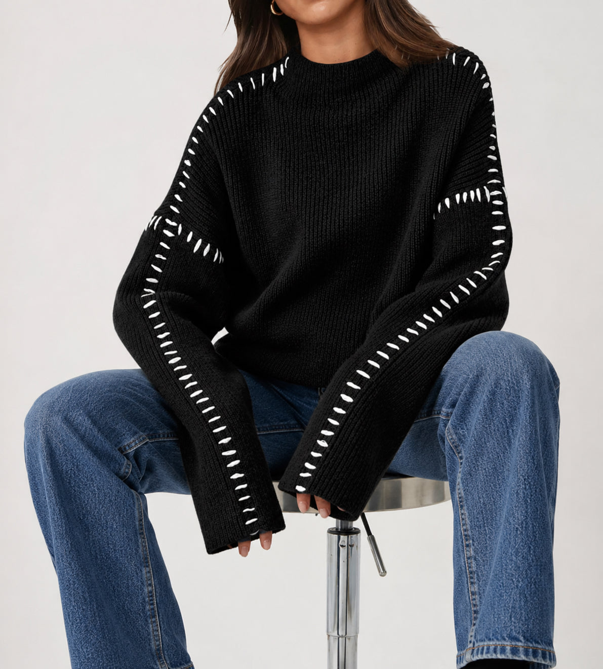 A person seated on a stool wearing an oversized black ribbed knit sweater with white stitched detailing along the sleeves and shoulders, paired with blue jeans, against a plain light background.