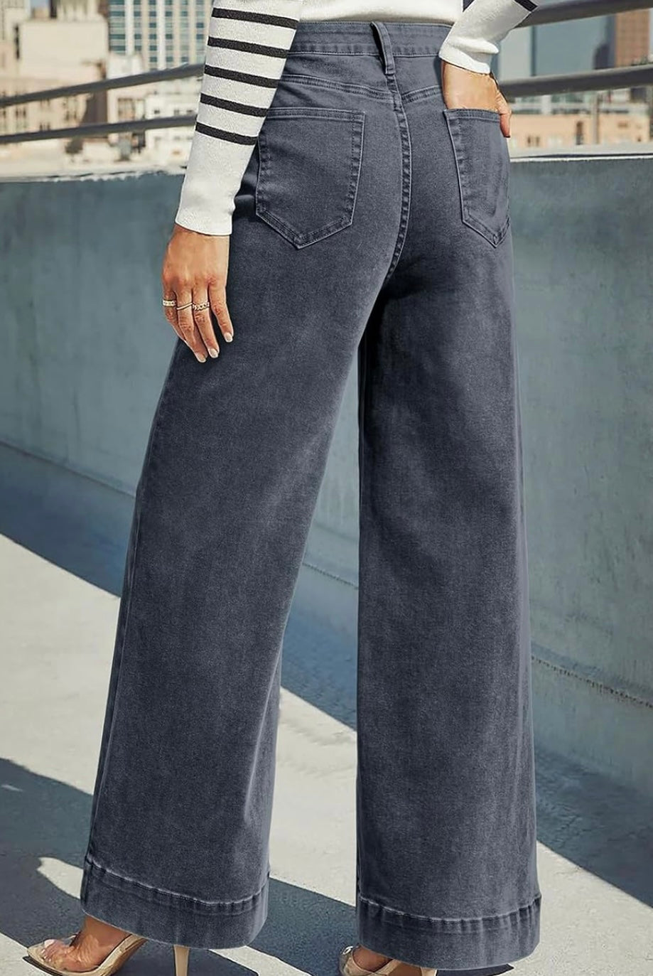 A person wearing dark gray wide-leg denim jeans, shown from the back in an outdoor urban setting with a concrete wall and buildings in the background.