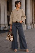 A woman stands outdoors in front of large stone columns, wearing a beige button-front cardigan, wide-leg dark gray jeans, and clear heeled sandals, while carrying a small cow-print handbag.
