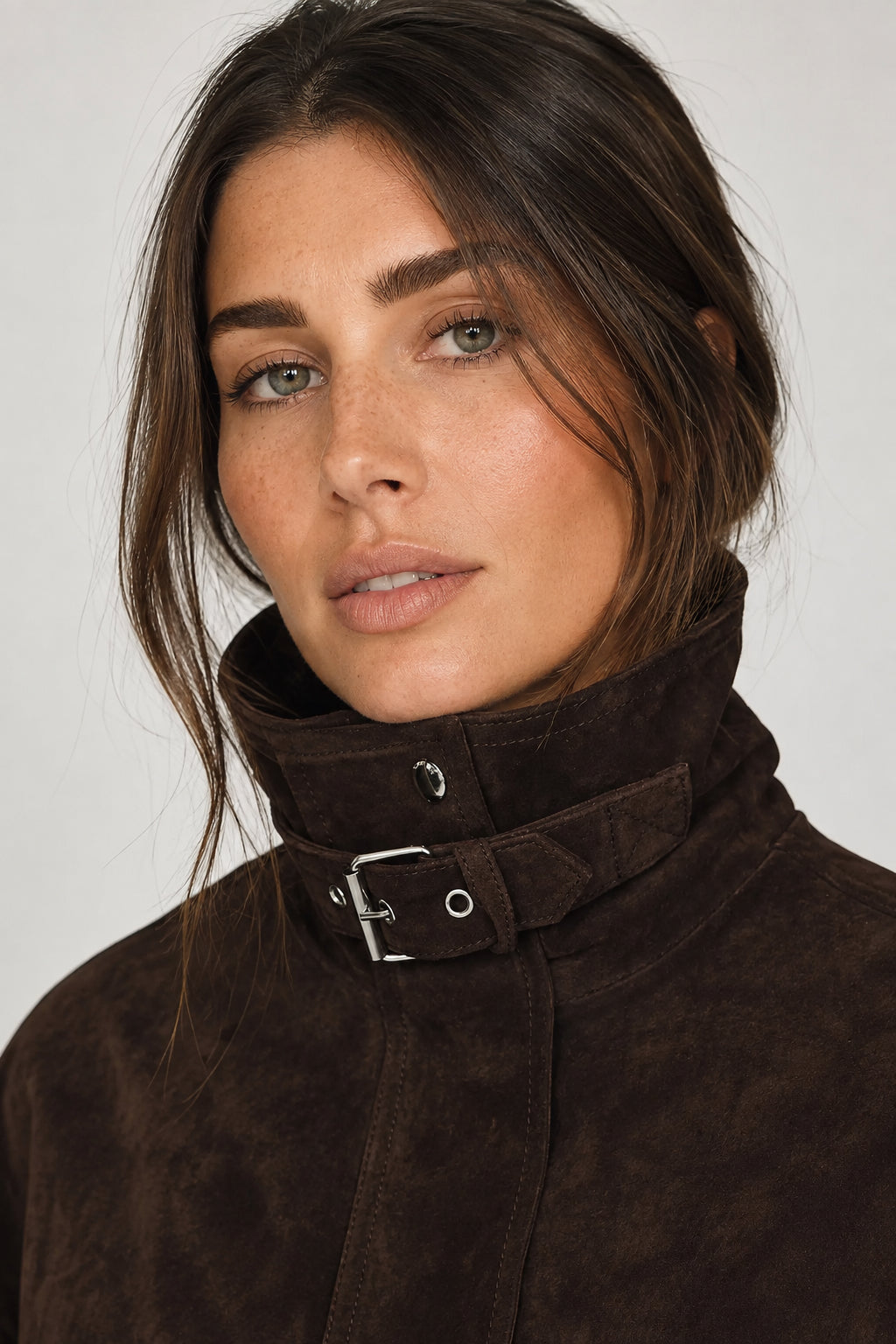 Close-up of a woman wearing a dark brown suede-like high-neck jacket with a wide collar and silver buckle detail, against a light gray background.