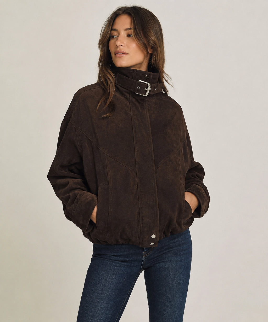 A woman wearing a dark brown suede jacket with a high collar and silver buckle detail, paired with dark blue jeans, against a plain light gray background.