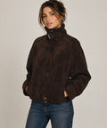 A woman wearing a dark brown suede jacket with a high collar and buckle detail, paired with blue jeans, photographed against a neutral light gray background.