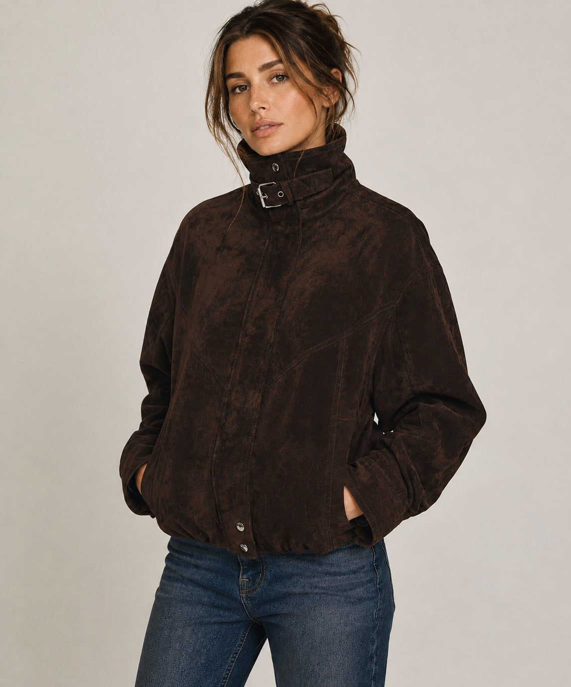 A woman wearing a dark brown suede jacket with a high collar and buckle detail, paired with blue jeans, photographed against a neutral light gray background.