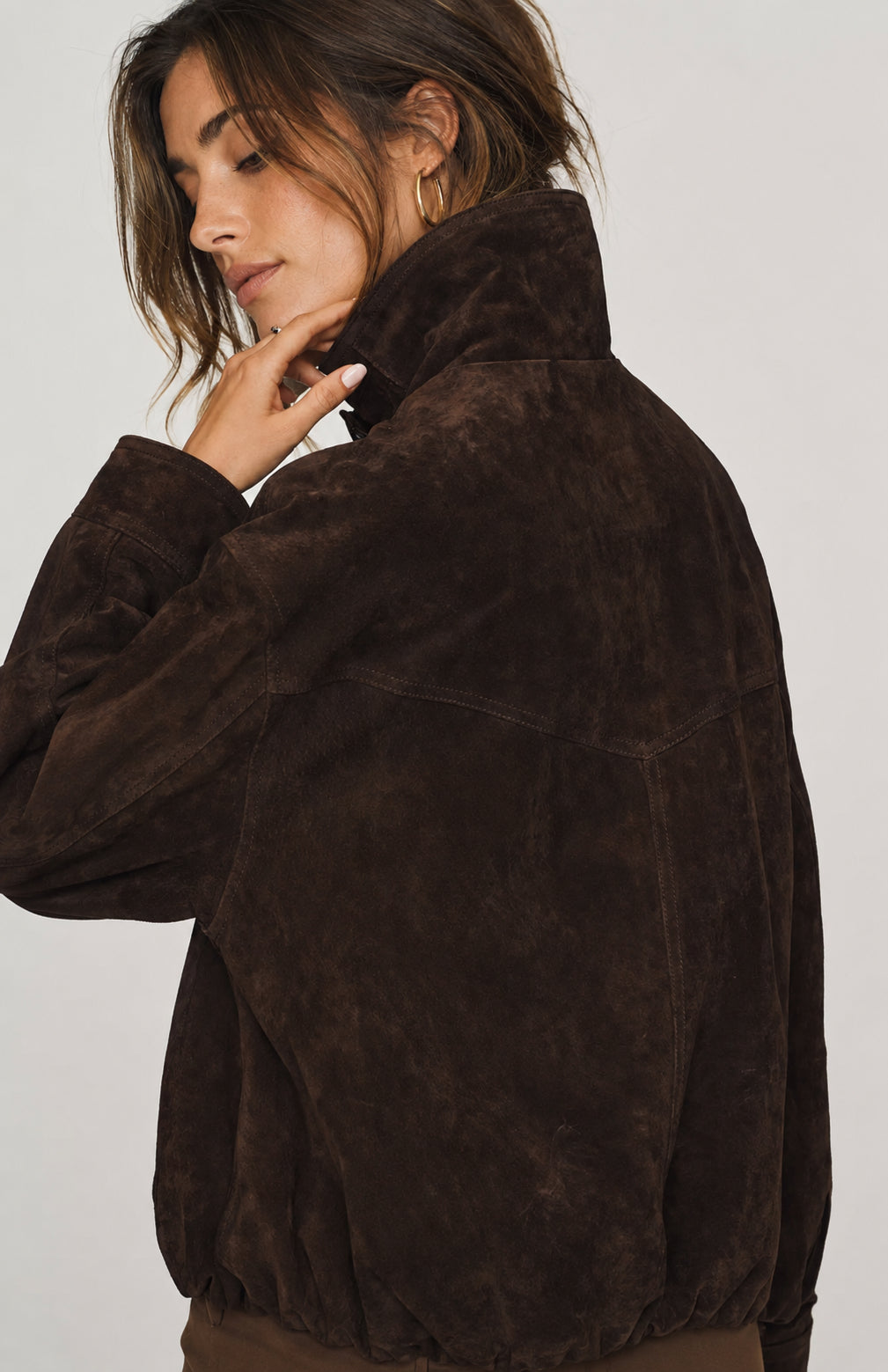 A woman wearing a dark brown suede jacket, shown from a side-back angle against a light gray studio background.