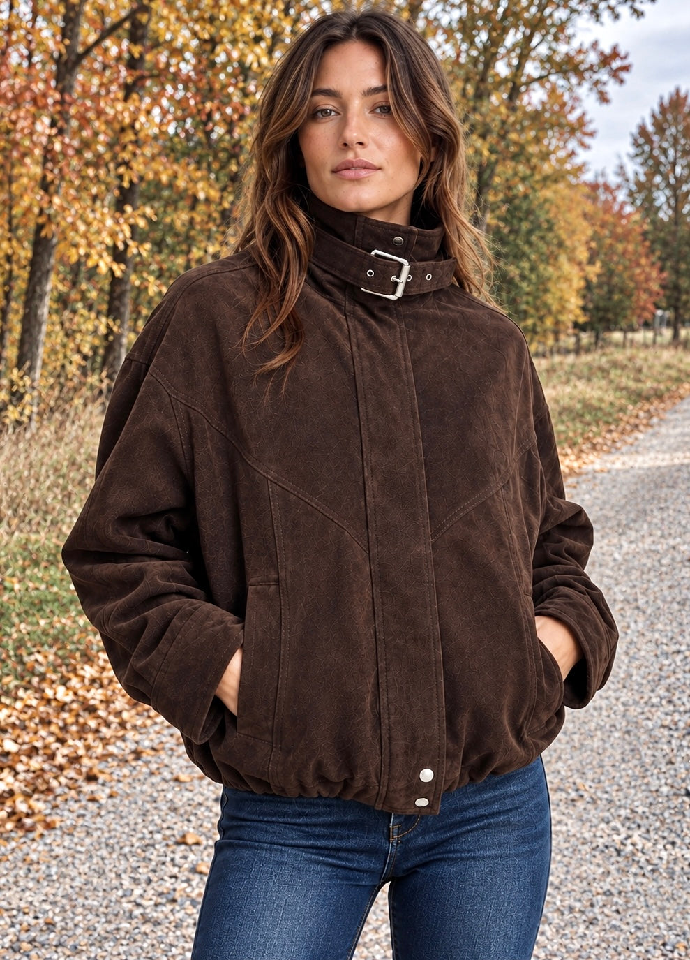 A woman wearing a dark brown suede jacket with a high collar and silver buckle, paired with blue jeans, standing on a gravel path surrounded by autumn trees.