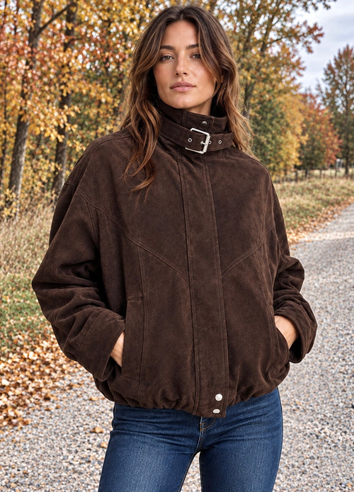 A woman wearing a dark brown suede jacket with a high collar and silver buckle, paired with blue jeans, standing on a gravel path surrounded by autumn trees.