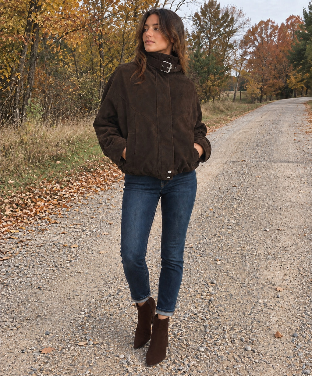 A woman stands on a gravel road in an autumn outdoor setting, wearing a dark brown suede jacket with a high collar and a silver buckle detail, blue jeans, and brown ankle boots.