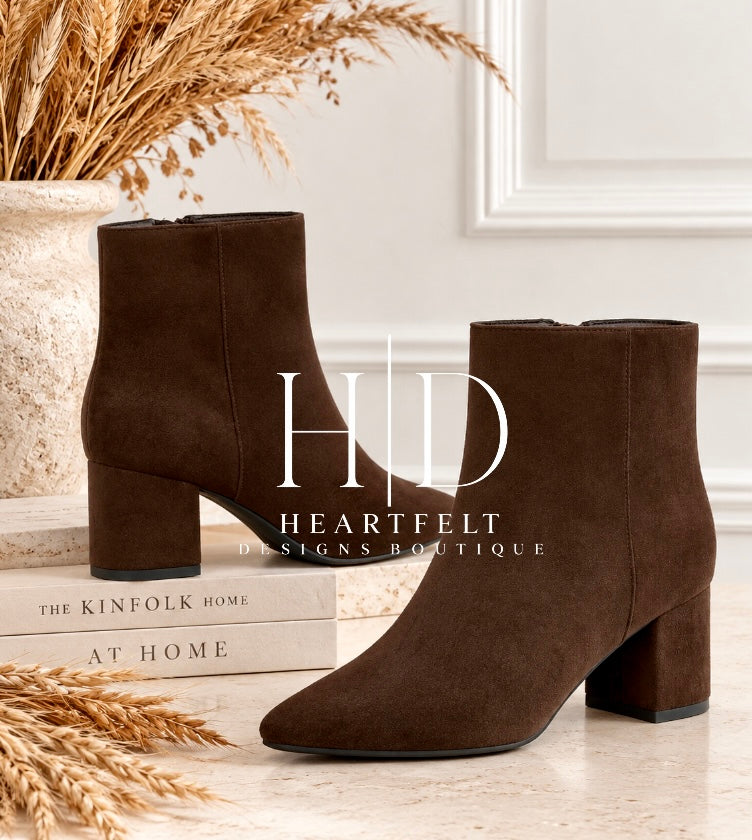 A pair of dark brown suede ankle boots with block heels, styled on a light stone surface with decorative books, dried grass, and a vase in a bright indoor setting. A large semi-transparent 'HID Heartfelt Designs Boutique' watermark is overlaid across the center.