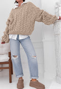 A person wearing an oversized beige cable-knit sweater, light-wash ripped jeans, and brown slip-on shoes, standing indoors near a white mantel and chair.