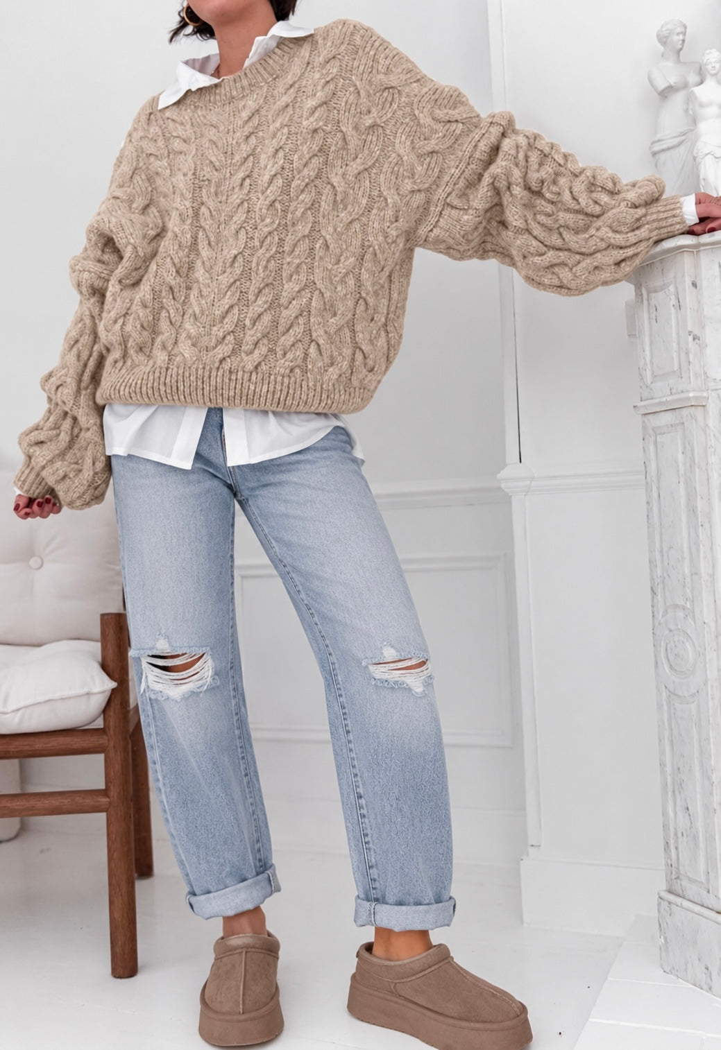A person wearing an oversized beige cable-knit sweater, light-wash ripped jeans, and brown slip-on shoes, standing indoors near a white mantel and chair.