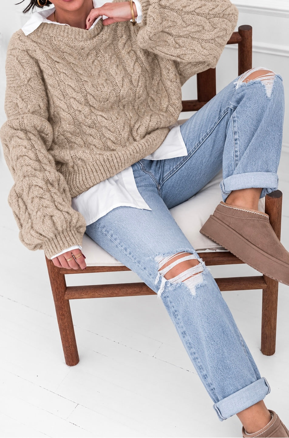 A person seated on a wooden chair wearing a chunky cable-knit beige sweater, light blue ripped jeans, a white shirt, and taupe slip-on shoes in a bright indoor setting.