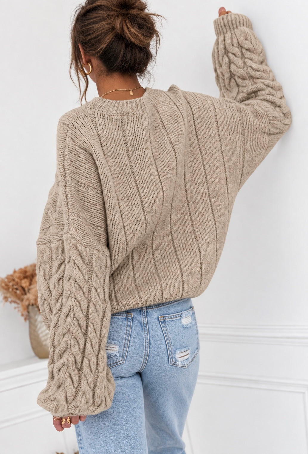 A woman shown from the back wearing a chunky cable-knit sweater in a warm taupe-beige color, paired with light-wash blue jeans, standing against a white wall.