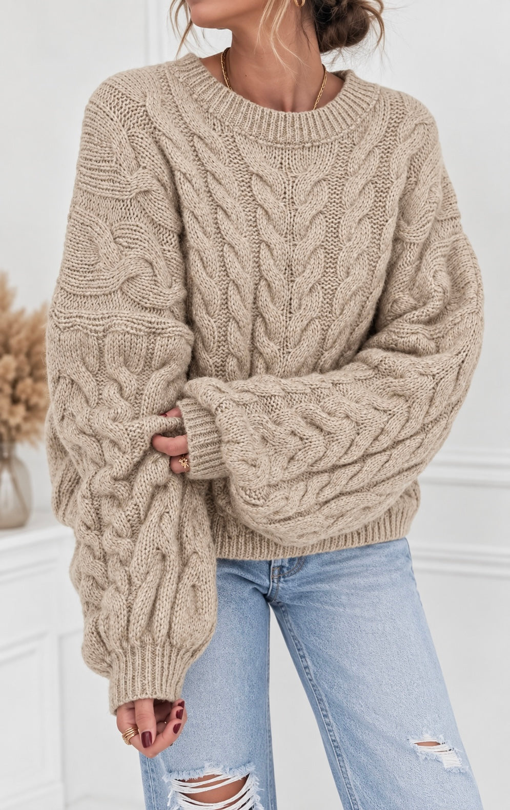A woman wearing a chunky cable-knit crewneck sweater in a warm beige color, paired with light-wash distressed jeans.