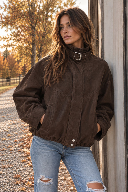 A woman wearing a dark brown corduroy jacket with a high collar and silver buckle detail, paired with light blue ripped jeans, standing outdoors next to a wooden wall.