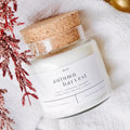 A white soy candle in a clear glass jar with a cork lid, labeled 'autumn harvest', styled on a white textured fabric with red dried botanicals.