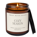 An amber glass jar candle with a lit wick and a black lid leaning against the right side, on a white background. The front label reads 'SWEET WATER DECOR' and 'COZY SEASON'.
