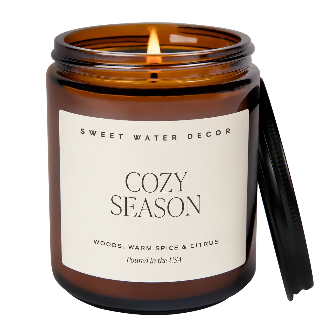 An amber glass jar candle with a lit wick and a black lid leaning against the right side, on a white background. The front label reads 'SWEET WATER DECOR' and 'COZY SEASON'.