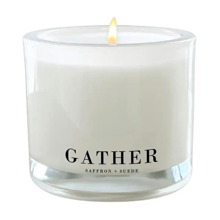 A lit white scented candle in a clear glass jar with the word GATHER printed in black on the front, plus smaller text reading SAFFRON + SUEDE.