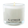 A lit white scented candle in a clear glass jar with the word GATHER printed in black on the front, plus smaller text reading SAFFRON + SUEDE.